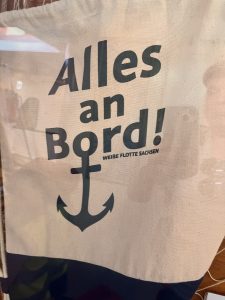 Ein Stoffbanner mit der deutschen Aufschrift „Alles an Bord!“ und einer Anker-Grafik. In kleinerer Schrift steht „WEISSE FLOTTE SACHSEN“. Das Design lässt auf ein nautisches oder maritimes Thema schließen.