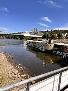 Ein Raddampfer liegt am Flussufer vor Anker, im Hintergrund sind eine nahegelegene Brücke, Bäume und Gebäude zu sehen, über ihnen erstreckt sich ein blauer Himmel mit vereinzelten Wolken.