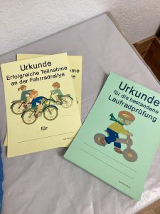 Auf einem Tisch liegen drei deutsche Urkunden: zwei gelbe für eine Fahrradrallye und eine grüne für eine Laufradprüfung; auf jeder sind Kinder abgebildet, die Fahrrad fahren.